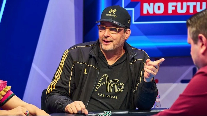 Phil Hellmuth, poker legend with 17 WSOP bracelets, at the No Future poker table on a streamed show, wearing a black Luxon Pay cap, glasses, and black Aria-branded jacket, pointing emphatically while talking during high-stakes play.