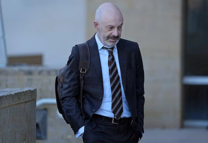 Tom Goldstein, prominent Supreme Court litigator and SCOTUSblog founder, arriving at court in a dark suit, white shirt, striped tie, and brown leather backpack, bald head and beard, walking outdoors with a thoughtful expression near a stone building.