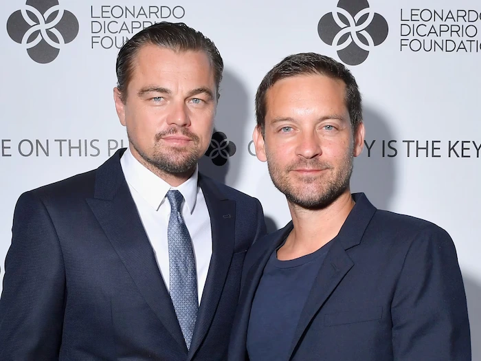 Tobey Maguire and Leonardo DiCaprio posing together on the red carpet at a Leonardo DiCaprio Foundation event, both in dark suits with Tobey in a blue tie and Leo in a gray one, standing in front of the foundation logo backdrop.