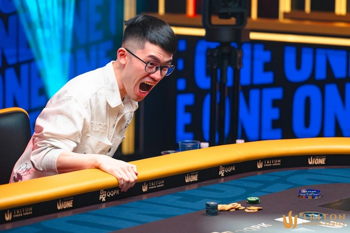 Zhou Yinan, Chinese high-stakes poker pro, leaning over the Triton Poker table in excitement, mouth open in celebration, wearing glasses and light shirt, arms on the yellow rail with Triton and UJONE branding, chips and cards visible on the blue felt, dramatic blue-orange lighting and "TRITON ONE" backdrop.