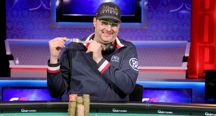 Phil Hellmuth, 17-time WSOP bracelet winner, grinning and holding up a Bitcoin-branded poker chip at the GGPoker high-stakes table, wearing a black Bitcoin cap, green-tinted glasses, navy jacket with Aria and Bitcoin patches, headphones around neck, with chip stacks and blue-lit studio backdrop.