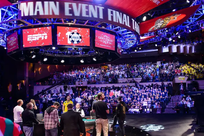 WSOP Main Event final table stage setup in a packed arena, featuring a massive overhead circular LED screen displaying "MAIN EVENT FINAL TABLE" with ESPN, 10 Poker Chip, and Gentleman Jack branding, bright spotlights, full crowd in stands, and players/staff at the green felt table below.