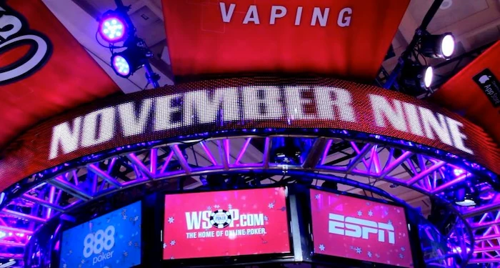 WSOP November Nine final table stage setup with illuminated "NOVEMBER NINE" LED sign overhead, WSOP.com and ESPN screens, 888 Poker branding, blue-purple lighting, red banners, and Vaping sponsor in the packed Las Vegas arena.