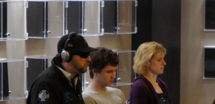 Phil Hellmuth in a black cap and headphones at a poker tournament with his wife Kathy Sanborn watching from the rail